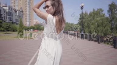 可爱的年轻女孩戴着太阳镜和一件白色的长夏时装走在户外。 一个漂亮女人的闲情逸致